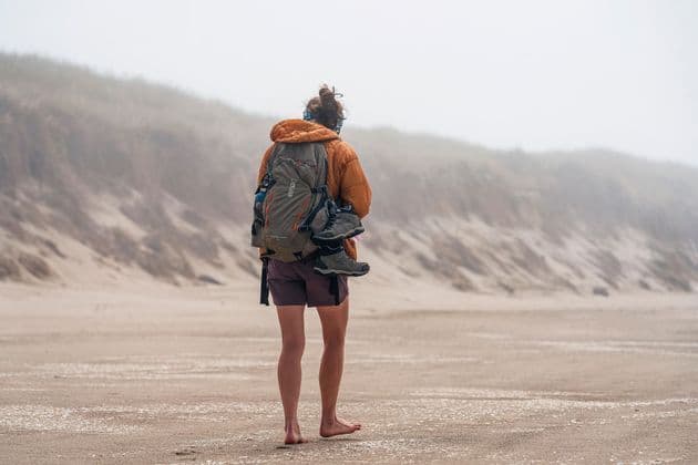 Una persona con mochila y chaqueta naranja camina descalza por una playa de arena en un día nublado.