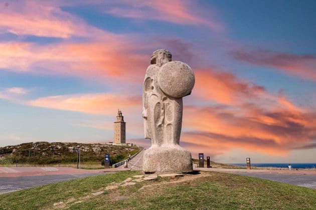 Una estatua de piedra de un guerrero con escudo se alza en una colina cubierta de hierba, con una torre de piedra al fondo contra un cielo de atardecer colorido.