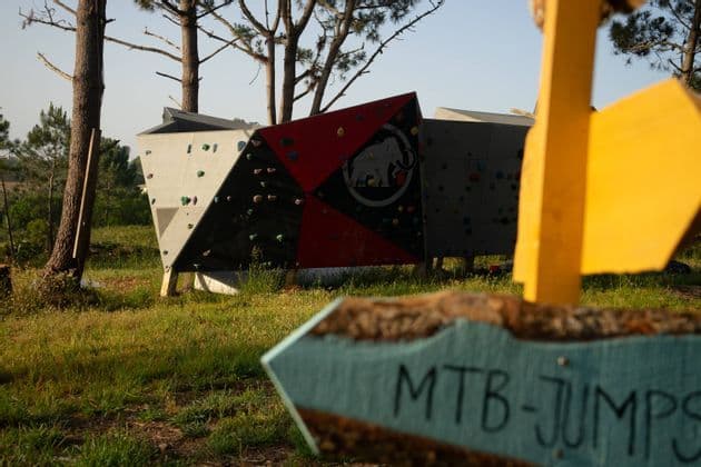 Un colorato masso da arrampicata all'aperto si trova in un parco erboso, con un cartello di legno con la scritta 'MTB-JUMPS' in primo piano sfocato.