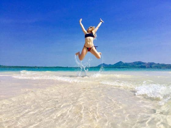 A woman in a bikini jumps out of the shallow, clear water at a tropical beach, with her arms raised towards the blue sky.