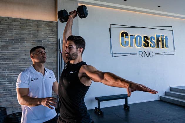 Un uomo esegue un sollevamento manubrio a braccio singolo sopra la testa mentre un allenatore lo osserva in una palestra CrossFit.