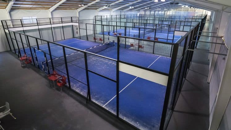 Vista dall'alto di campi da padel blu vuoti con pareti in vetro disposti in file all'interno di una grande struttura sportiva al coperto.