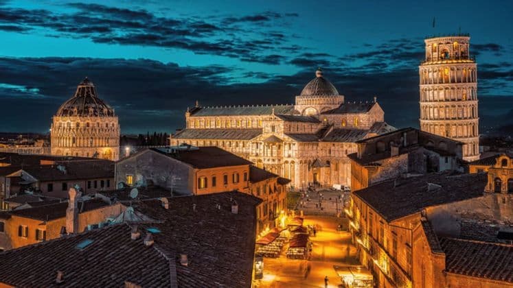 Veduta dall'alto di Torre Pendente, cattedrale e battistero illuminati al crepuscolo, sopra i tetti di una città storica.