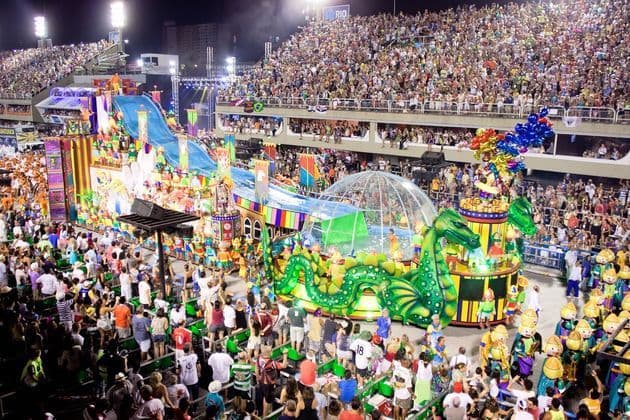 Un défilé coloré avec des chars élaborés, incluant un grand dragon vert, traverse un stade bondé de spectateurs la nuit.