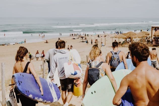 Un viaje en grupo de WeRoad visto desde atrás, llevando tablas de surf y caminando hacia el océano en una playa concurrida.