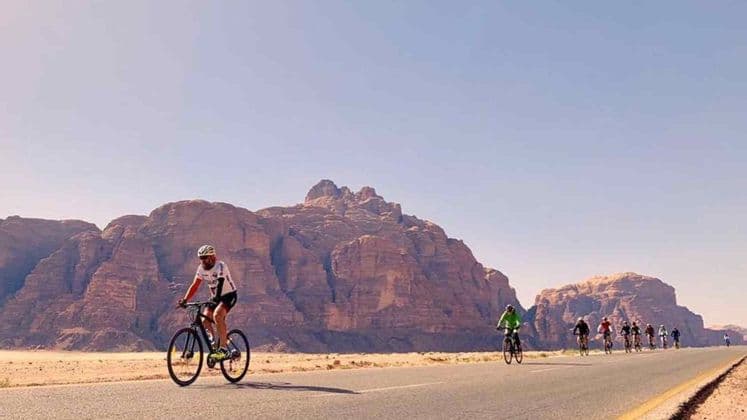 Un viaggio di gruppo WeRoad con persone in bicicletta su una strada asfaltata attraverso un paesaggio desertico con grandi formazioni rocciose sullo sfondo.