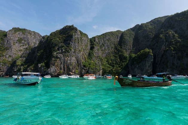 Barche tradizionali in legno con nastri colorati sono ormeggiate su una spiaggia sabbiosa in acqua turchese, con una grande isola carsica calcarea sullo sfondo.