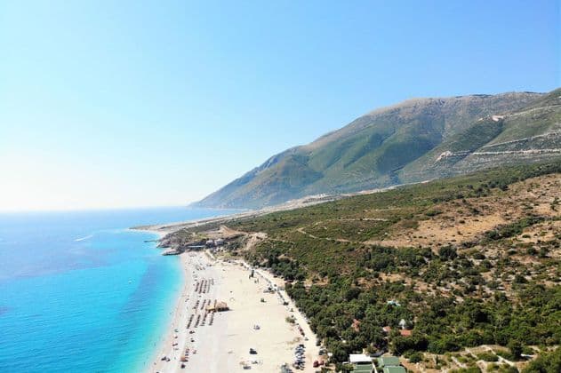 Eine Luftaufnahme eines langen Sandstrandes mit türkisfarbenem Wasser, gelegen neben einem großen grünen Berg unter klarem blauem Himmel.