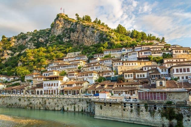 Un conjunto de casas blancas históricas con tejados de tejas agrupadas en una ladera verde que domina un río, con una fortaleza en la cima.