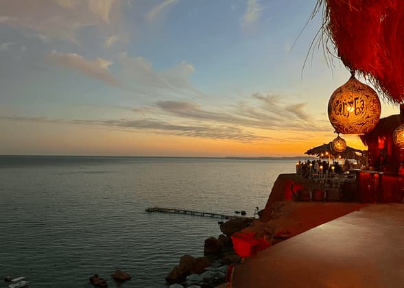 Vista da un ristorante sulla scogliera con affaccio sul mare durante un tramonto colorato, con una lanterna ornata in primo piano.