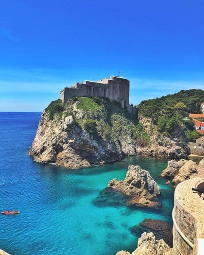 Una fortezza in pietra si erge su una scogliera rocciosa e verde che domina un mare turchese con due kayak rossi che remano nell'acqua sottostante.