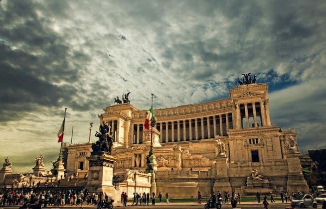 Un gran monumento clásico con columnas, estatuas y banderas italianas se alza bajo un cielo oscuro y nublado mientras la gente camina.