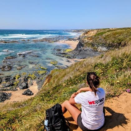 Ein WeRoad Gruppenleiter sitzt mit Rucksack auf einer grasbewachsenen Klippe und blickt auf eine felsige Küste und das Meer.
