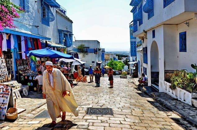 Un homme en robes traditionnelles marche le long d'une rue pavée ensoleillée bordée d'étals de marché et de bâtiments blancs et bleus.