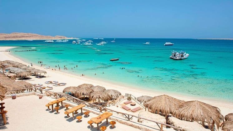 Una spiaggia di sabbia bianca con ombrelloni di paglia si affaccia su una baia dalle acque limpide e turchesi, con numerose barche ancorate al largo.