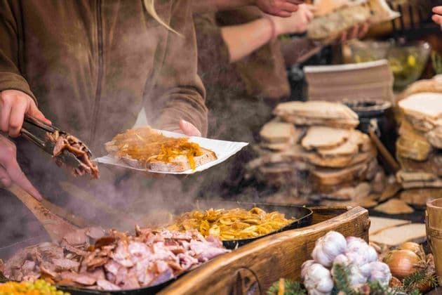 Un vendedor en un puesto de comida sirve carne y cebolla calientes de una sartén humeante sobre una rebanada de pan en un plato de papel.