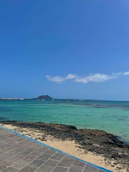 Un paseo pavimentado junto a una costa rocosa con vistas a una bahía turquesa con veleros y una isla lejana bajo un cielo azul claro.