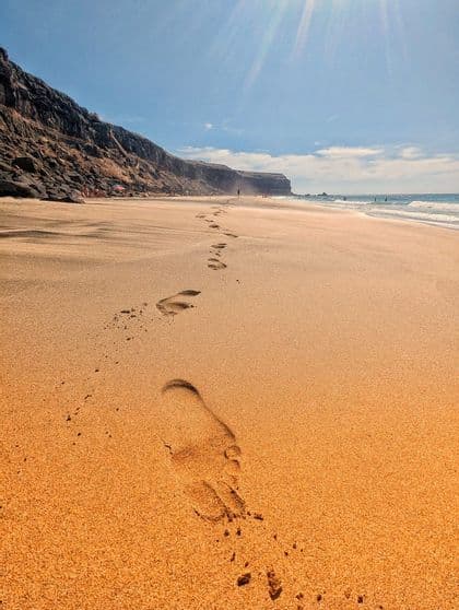 Primer plano de huellas que descienden por una playa de arena dorada, con un acantilado rocoso a un lado y el océano al otro.