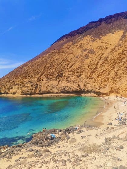 Una cala de playa apartada con agua turquesa al pie de un acantilado grande, empinado y de color ocre bajo un cielo azul claro.