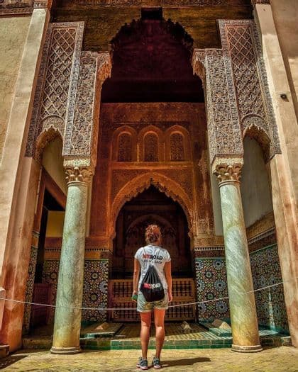 Una mujer con camiseta y mochila de WeRoad de pie mirando hacia un gran arco ornamentado con tallas detalladas y mosaicos de azulejos.