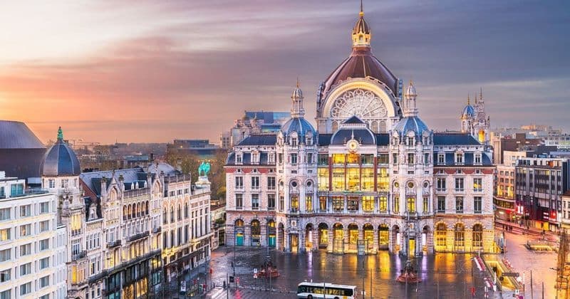 Un grand bâtiment historique avec un dôme, des fenêtres illuminées surplombant une place de ville humide au coucher du soleil.