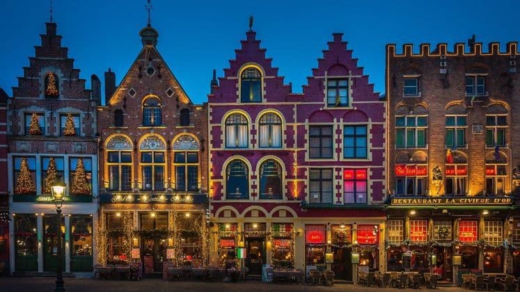 Une rangée de bâtiments historiques colorés aux pignons à redents, décorés de lumières de Noël et illuminés au crépuscule.