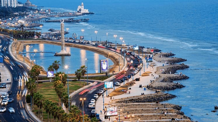 Vue aérienne d'une ville côtière au crépuscule, montrant une autoroute incurvée longeant la mer, avec un grand monument dans une lagune.