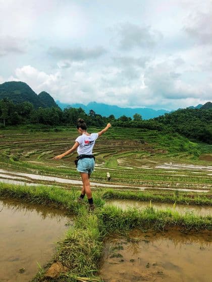 A woman on a WeRoad trip balances on a narrow, grassy path between terraced rice paddies, with mountains in the background.