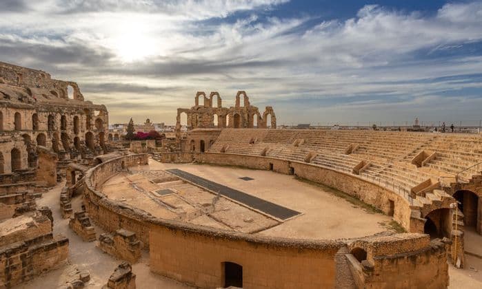 The vast ruins of a stone amphitheater, showing the central arena and tiered seating under a cloudy sky.