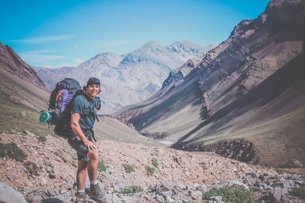A smiling hiker with a large backpack pauses on a rocky trail in a vast mountain valley.