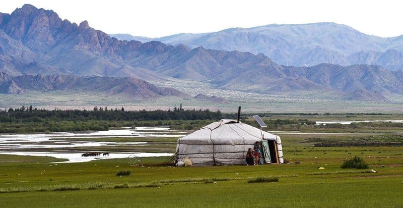 A family sits at the entrance of a traditional yurt in a vast green valley with a river and mountains in the background.