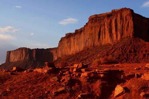 Large red rock formations are lit by warm sunlight, casting shadows across a rocky desert landscape under a blue sky.