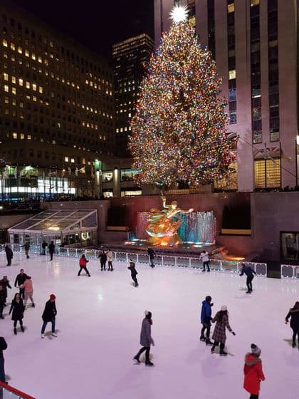 Personas patinando sobre hielo de noche en una pista al aire libre frente a un gran árbol de Navidad iluminado y altos edificios de la ciudad.