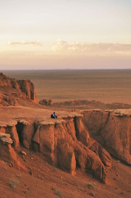 Eine Person mit einem Rucksack sitzt am Rande eines roten Felsencanyons und blickt während eines blassen Sonnenuntergangs über eine weite Wüstenebene.