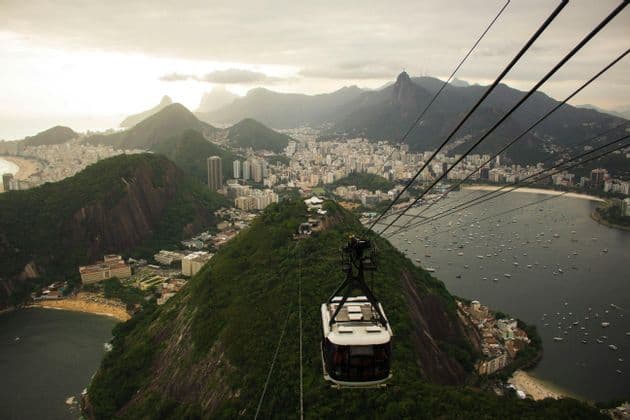 Vista aérea desde un teleférico sobre una ciudad costera, mostrando montañas verdes, una bahía con barcos y una estatua en un pico distante.