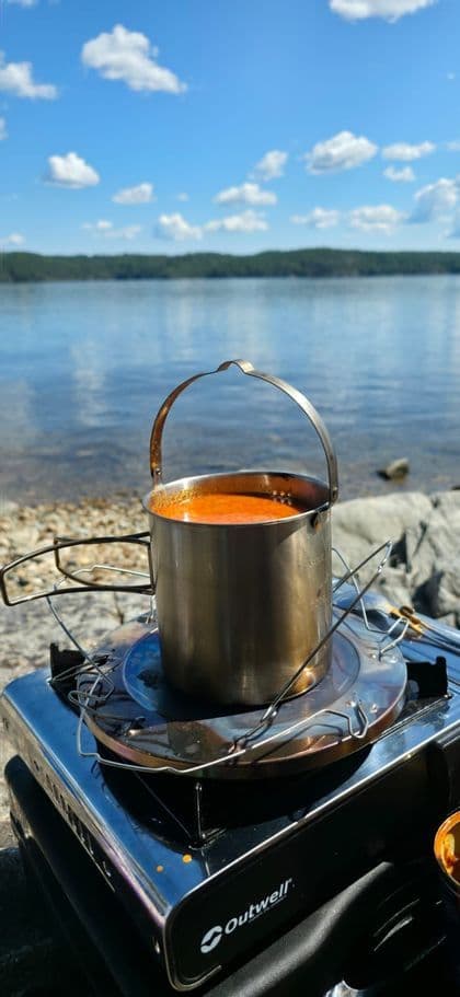 Ein Metalltopf mit orangefarbener Suppe kocht auf einem tragbaren Kocher am felsigen Ufer eines Sees unter blauem Himmel.