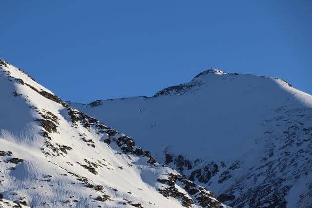 Picos montañosos nevados y soleados con formaciones rocosas oscuras resaltan contra un cielo azul claro