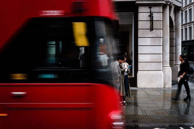 Un autobús rojo con desenfoque de movimiento pasa a toda velocidad junto a dos personas de pie en una acera mojada de la ciudad, mirando un libro.