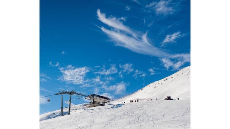 Una stazione della seggiovia di un comprensorio sciistico su un pendio montano innevato con sciatori e un gatto delle nevi sotto un cielo azzurro vivido.
