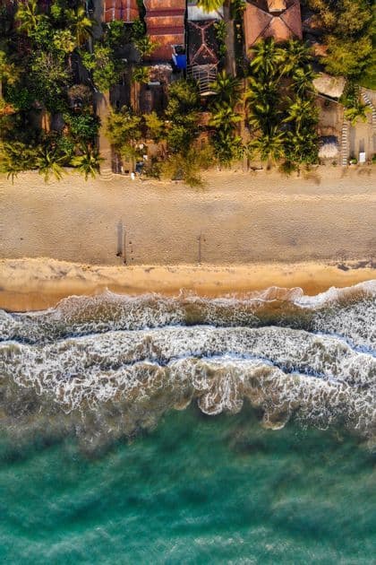 Vista aerea dall'alto di una spiaggia tropicale, con acqua turchese e onde spumose che si infrangono sulla sabbia vicino alle palme.