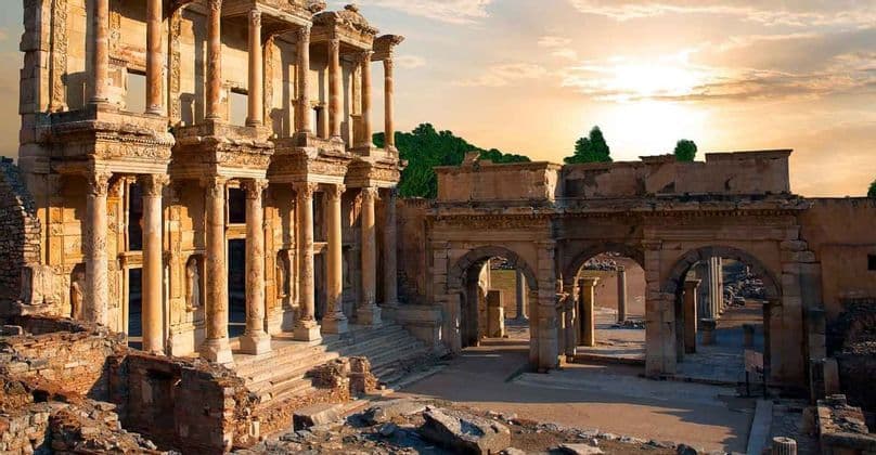 La façade d'une ancienne bibliothèque en pierre avec de hautes colonnes se dresse au milieu des ruines au coucher du soleil, illuminée par une lumière dorée.