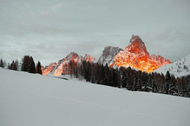 Le vette rocciose delle montagne sono illuminate dalla luce arancione del tramonto su un paesaggio innevato con una fitta pineta.