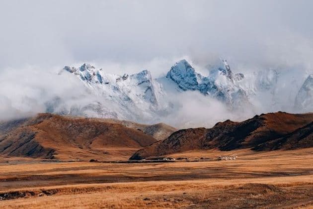 Picos de montañas nevadas se elevan entre las nubes sobre una vasta estepa dorada con un pequeño asentamiento en la distancia.