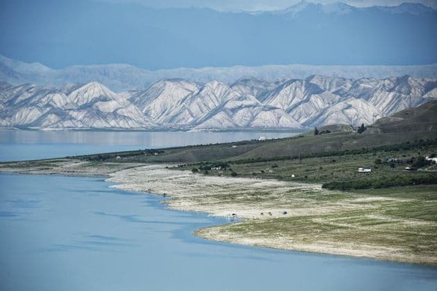 Un vasto lago blu si curva lungo una costa di verdi colline, con strati di catene montuose bianche e blu sullo sfondo.