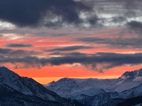 Un tramonto colorato con sfumature arancioni e rosa sotto nuvole scure illumina una catena montuosa innevata.
