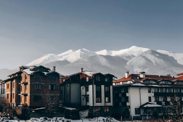 Una hilera de edificios alpinos con tejados nevados se asienta frente a una gran cordillera cubierta de nieve bajo un cielo despejado.