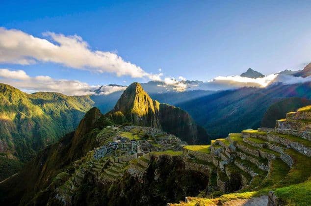 A panoramic view of ancient stone ruins and terraces on a green mountainside with a prominent peak lit by sunlight.