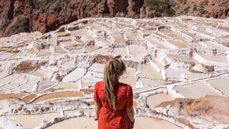 A woman with blonde dreadlocks in a red top, seen from behind, looking out over terraced salt evaporation ponds.