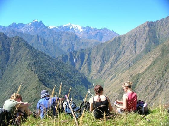 A WeRoad group trip taking a break on a grassy mountainside, admiring a view of a mountain valley and distant snowy peaks.