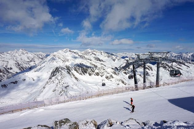 Deux personnes skient sur une piste à côté d'un télésiège, avec une vue panoramique sur des montagnes enneigées sous un ciel bleu.
